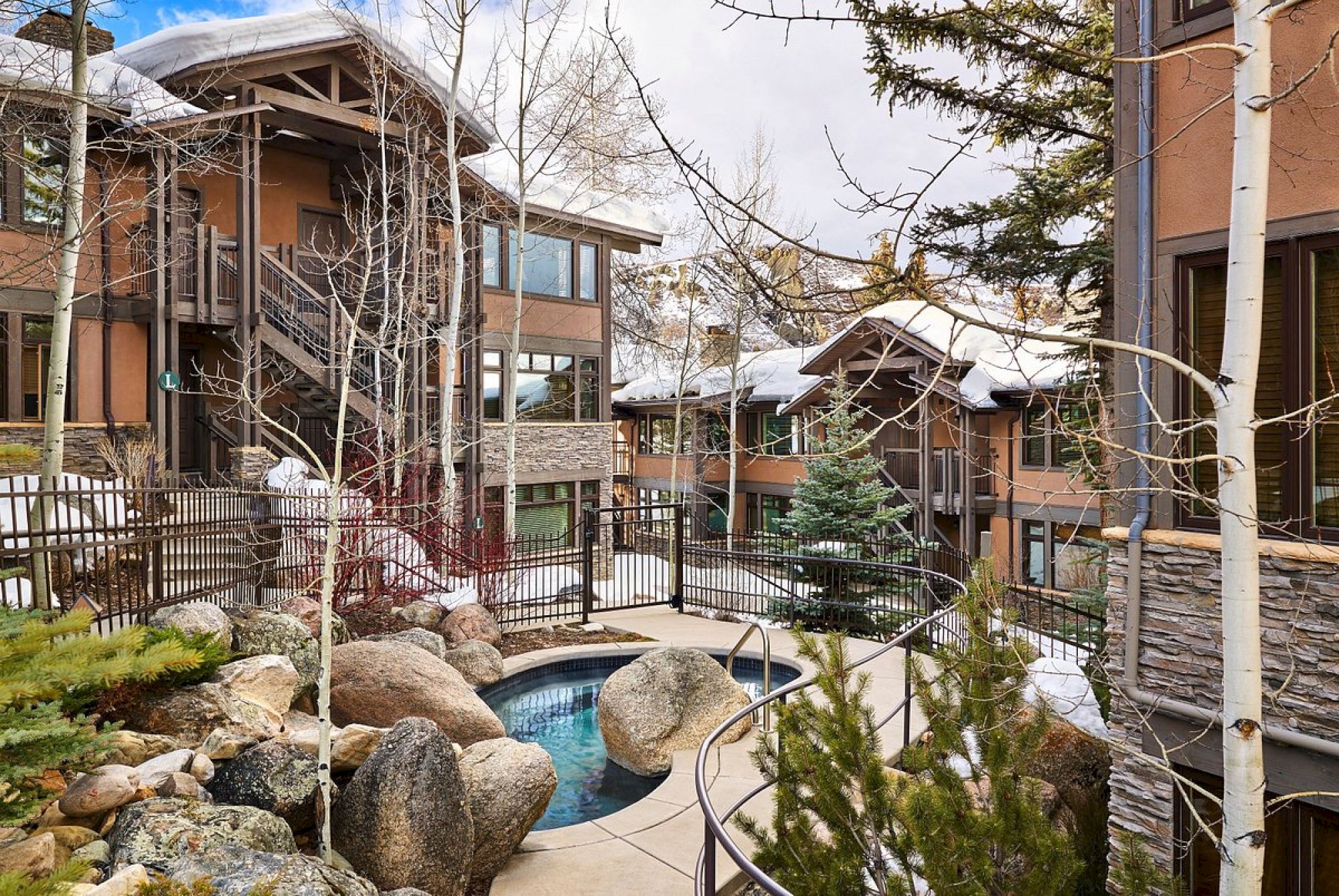 A scenic residential area featuring rustic-style buildings, trees, rocks, a small hot tub or pool, and snow-covered roofs and landscape.