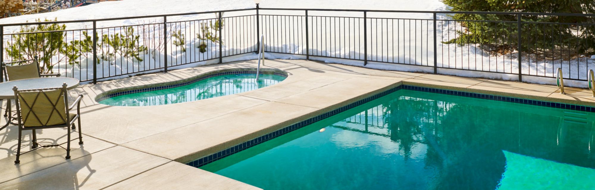 The image shows a swimming pool and hot tub surrounded by a fence during winter, with snow on the ground outside the pool area.