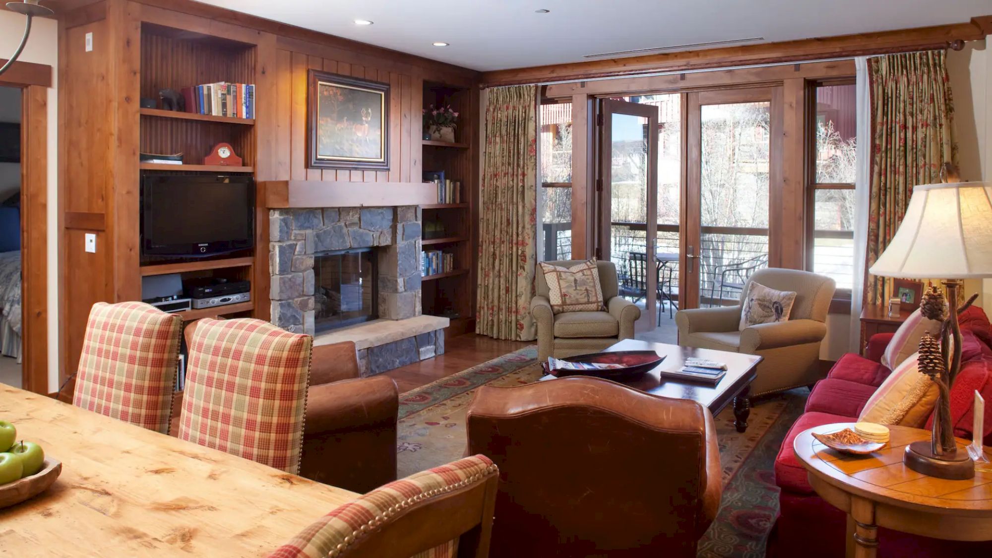 The image shows a cozy living room with wooden accents, a stone fireplace, comfortable seating, a television, and large windows overlooking a patio.