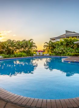 A serene outdoor swimming pool surrounded by palm trees and lounge chairs at sunset, reflecting a calm, relaxing atmosphere.