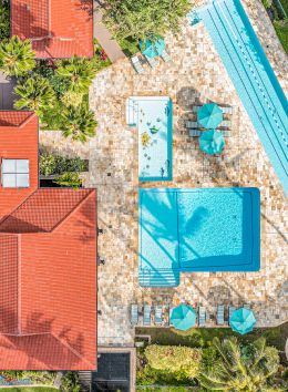 Aerial view of a property with a pool, small outdoor seating areas under umbrellas, and a red-roofed building surrounded by greenery ending the sentence.