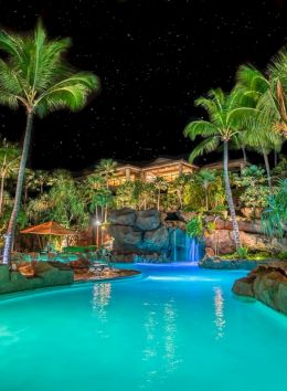 A nighttime scene of a tropical pool lit with blue lights, surrounded by palm trees and rocky landscaping under a starry sky.