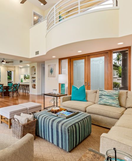 A spacious living room with beige sofas, turquoise accents, striped ottoman, and light wooden flooring, opens into a dining area and an upper balcony.