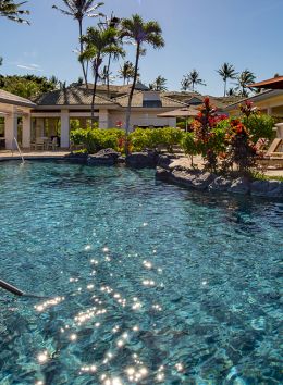 The image shows a serene outdoor swimming pool surrounded by lounge chairs, umbrellas, palm trees, and cabanas on a sunny day.