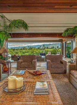 A spacious living room with large sofas, palm plants, candles, and books on a central coffee table, opening to a scenic outdoor view.