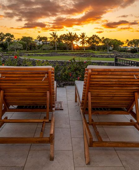 Two wooden lounge chairs face a scenic sunset view, with a lush garden and trees in the background. The setting is serene and picturesque.