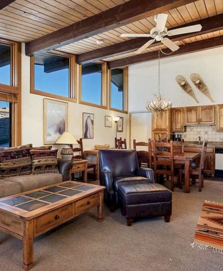 A cozy living room with rustic decor, featuring a stone fireplace, wooden furniture, a leather armchair, a sofa, and a kitchen area in the background.