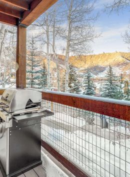 The image shows a snow-covered balcony with a grill, surrounded by trees and a scenic mountain view in the background.