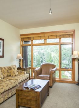 A cozy living room with a floral sofa, leather armchair, wooden coffee table, lamps, wall art, large windows, stone fireplace, and wall-mounted TV.