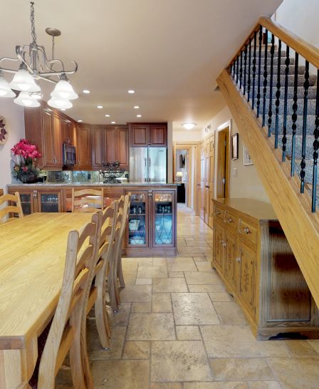 The image shows a dining area with a long wooden table, chairs, chandelier, decorative items, and a staircase leading to an upper floor.