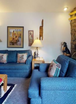 A cozy living room with blue sofas, wooden coffee table, fireplace, wall art, and decorative items on shelves. The room is warmly lit.