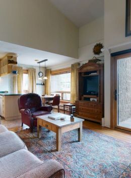 A cozy living room with a beige sofa, a wooden coffee table, a TV, and a balcony view. The space leads to a kitchen and dining area.