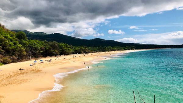 A scenic beach features turquoise waters, golden sand, and people relaxing under partly cloudy skies, with lush green hills in the background.