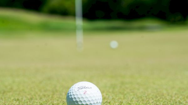 A close-up of a golf ball on a green field with another ball and a flag in the background, under a clear sky.