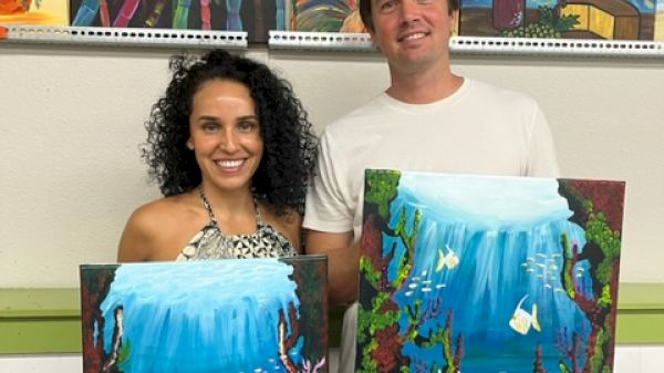 Two people are standing and holding paintings of underwater scenes, smiling. More artwork is displayed on the wall behind them.