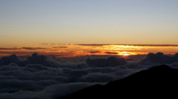 The image shows a serene sunrise or sunset over a sea of clouds with a silhouetted mountain in the foreground and a clear sky above.