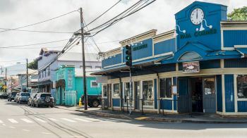 A street view featuring colorful buildings, a clock on the blue building, parked cars, and power lines under a cloudy sky.