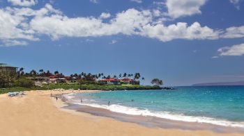 A sandy beach with turquoise waters, a few people swimming, buildings in the background, and palm trees under a partly cloudy sky.