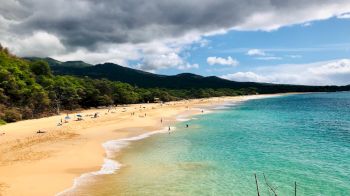 A pristine beach with golden sand and crystal-clear turquoise water, surrounded by lush greenery and backed by cloudy skies.