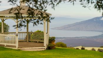 A white gazebo stands on a grassy hill with a scenic view of a body of water and distant mountains, framed by leaves.