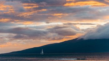 A serene sunset over the ocean with a sailboat in the distance, and a cloud-covered mountainous island in the background.