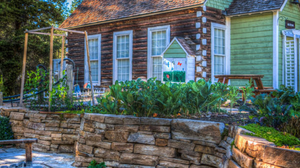 A rustic house with a garden, surrounded by trees, stone walls, and a paved pathway leading to a seating area.