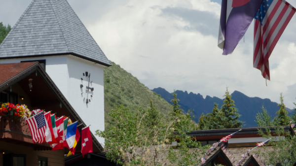 The image shows a charming village with alpine-style buildings adorned with various national flags, vibrant flowers, and the Colorado state and US flags.