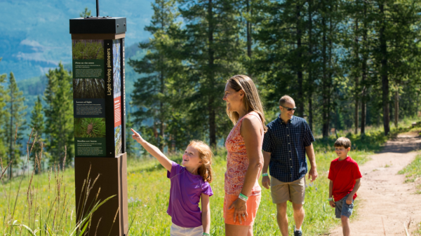 A family with two children walks on a forested trail, while the daughter points at an informational signpost with mountains visible in the background.