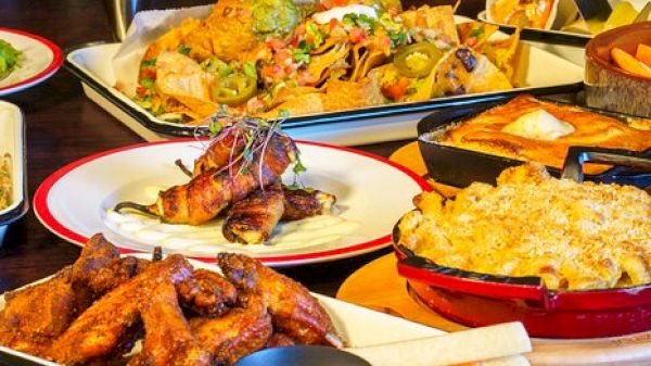 The image showcases a spread of dishes including nachos, chicken wings, macaroni and cheese, a baked dish, and assorted appetizers.