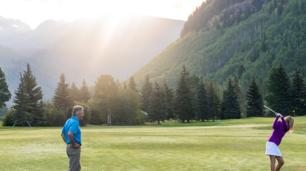 A person in a purple shirt swings a golf club while another person in a blue shirt watches, with a scenic backdrop of mountains and trees.