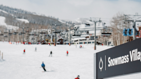 The image shows a ski slope with people skiing and snowboarding at Snowmass Village. The background features ski lifts and snow-covered buildings.