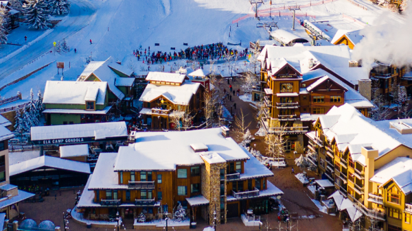 The image shows a snowy ski resort with buildings, snow-covered rooftops, ski slopes, and people gathered near the slopes in a mountainous area.