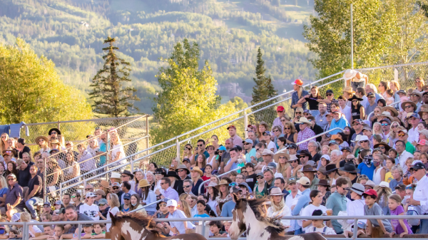 A large crowd watches a rodeo with three horses in an enclosure, with scenic mountains and trees in the background under a clear sky.