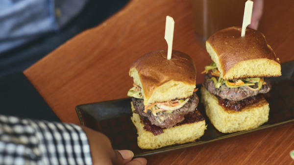 A person holding a glass of beer, while another person is placing a plate with two sliders on a wooden table, next to some small plants.