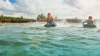 Two people are riding bodyboards in the ocean near a sandy beach with palm trees and cloudy skies.