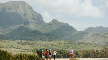 A group of people are standing on a rocky outcrop with a lush mountainous landscape and a cloudy sky in the background, all seeming to be outdoors.