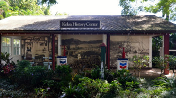 A building with a sign that reads "Koloa History Center," featuring Chevron gas pumps, historical photos, and lush greenery in the foreground.