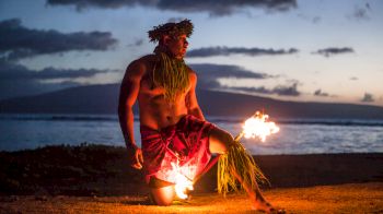 A person in traditional attire performs a fire dance on a beach at sunset, with the ocean and mountains visible in the background, ending the sentence.