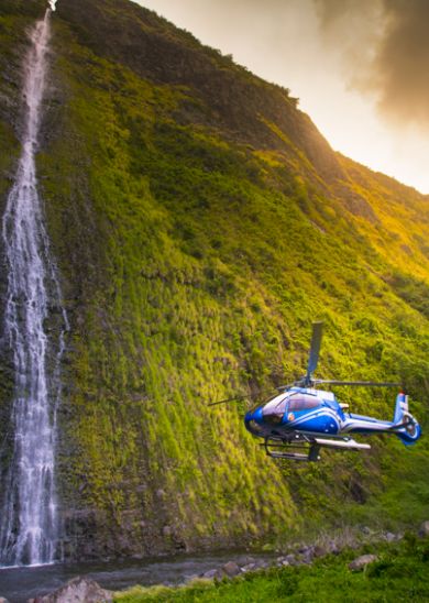 A blue helicopter flies near a tall, narrow waterfall cascading down a lush, green mountainside under a cloudy sky at sunset.