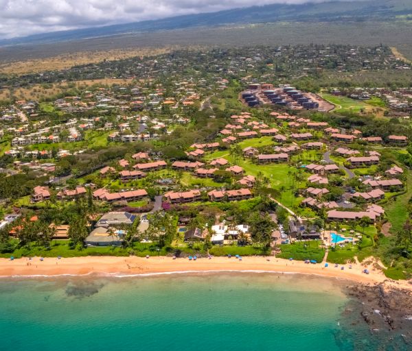 Aerial view of a coastal community with green spaces, residential buildings, and a beach with teal-colored water and some waves along the shore.