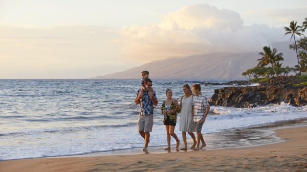 A group of five people walks along a sandy beach near the ocean at sunset, with mountains and palm trees in the background.