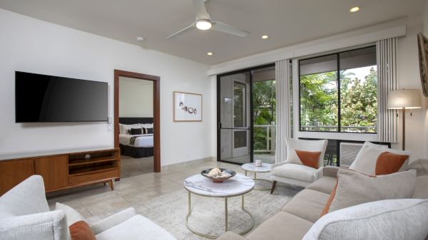 A modern living room with a TV, beige couches, a round coffee table, and large windows overlooking greenery. A doorway leads to a bedroom.