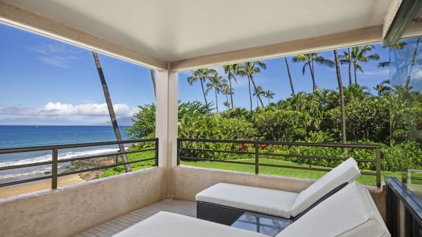 A relaxed balcony with two lounge chairs, providing a view of a tropical beach with palm trees, blue sky, and ocean waves on a sunny day.