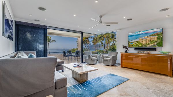 A modern living room features a sofa, coffee table, and large flat-screen TV. Sliding doors open to a balcony with a beach view, showcasing palm trees.