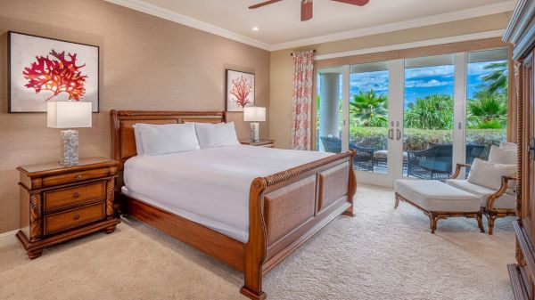 A cozy bedroom with a wooden bed, two nightstands with lamps, artwork, a ceiling fan, and glass doors leading to an outdoor area with lush greenery.