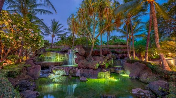 The image features a serene, tropical garden scene with a waterfall, lush greenery, rocks, and palm trees, beautifully lit at dusk.