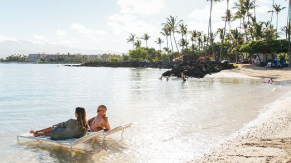 A woman and a child relax on a chair in shallow water at a sunny beach, with palm trees and people in the background.