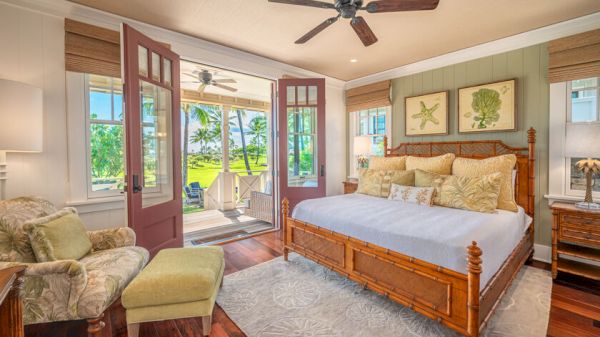 A cozy bedroom with a large bed, armchair, footrest, and two lamps, opens to an outdoor space with chairs and greenery visible through glass doors.