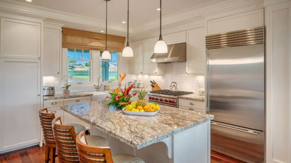 A modern kitchen featuring a large island with granite countertop, three stools, stainless steel appliances, and fresh flowers and fruit on the counter.