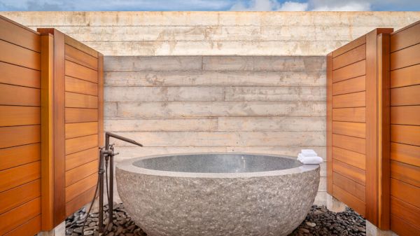 The image shows an outdoor stone bathtub with wooden privacy panels on either side, a faucet, and a stack of white towels against a concrete wall.